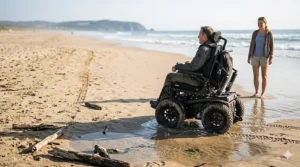 A specialized all terrain power wheelchair with wide tires parked on a sandy beach near the ocean.