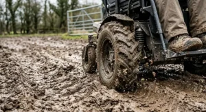 A close-up view of the rugged, deep-tread tires on an electric all-terrain wheelchair as it powerfully handles thick mud on a wet farm track.