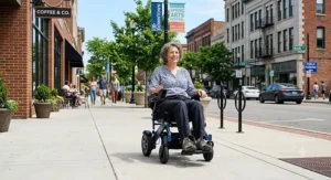 A person riding a travel power wheelchair smoothly over a paved public sidewalk in a sunny American downtown area.