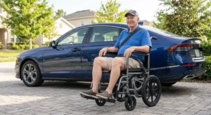 An elderly American man sitting in a portable wheelchair next to a parked car on a sunny day.