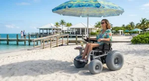A vacationer using an all-terrain beach wheelchair under a tropical beach umbrella enjoying a resort view.